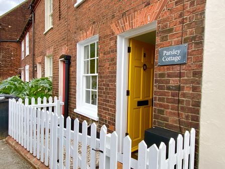 A brick exterior of a cottage with a white picket fence and a yellow door at Parsley Cottage Southwold