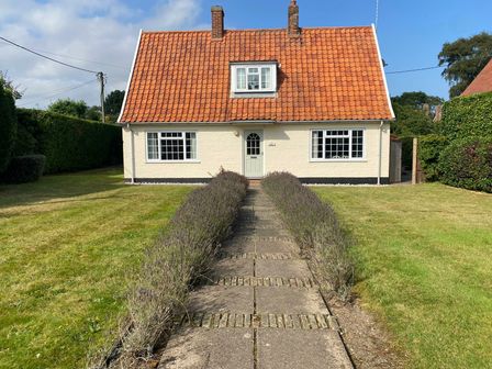 A house with a red tiled roof and white walls with a paved path and shrubs leading to the front door at Hoist Cottage Walberswick