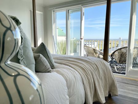 A bedroom with a bed, pillows, a blanket, sliding glass doors leading to a balcony with wicker chairs overlooking a landscape at The Dunes, Southwold