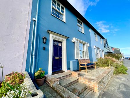 A blue painted house exterior with a dark blue door and a wooden bench on a paved porch at 11 Crabbe Street Aldeburgh