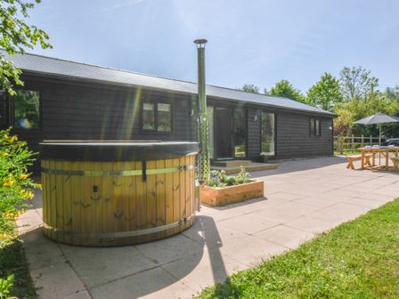 An outdoor patio with a wooden hot tub a flower planter a picnic table with an umbrella and a black-clad building at The Piglets in Newbourne Near Woodbridge