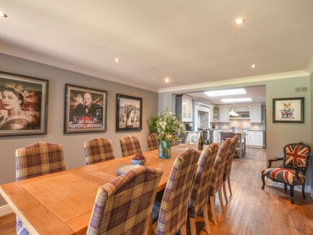 A dining room with a long wooden table and plaid chairs next to an open kitchen at Moat Barn in Hasketon Near Woodbridge