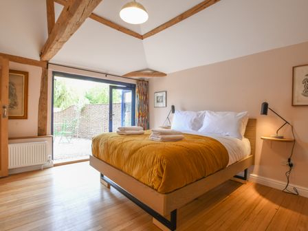 A bedroom with a wooden bed with yellow bedding and white pillows and folded towels by black lamps and glass doors leading outside at Onna's Stable Sotherton in Sotherton near Halesworth