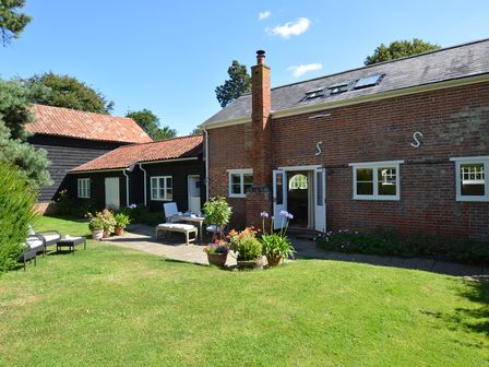 A garden with grass and potted plants next to a brick cottage with outdoor seating at Stable Cottage at the Grove Great Glemham Near Framlingham