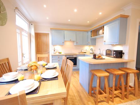 A kitchen with blue cabinets wooden stools a dining table set with plates glasses of orange juice and a bowl of fruit at Garden Cottage Southwold