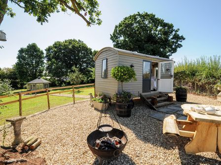A small wooden cabin with open doors on a gravel area with a fire pit and wooden table with cups and teapot at Orchard Retreat East Worlington near Witheridge