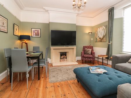 A living room with a dining table and grey chairs a tv above a fireplace an armchair and a blue ottoman at Spinners House in Dartmouth