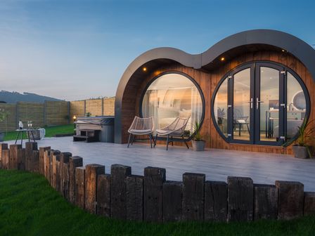 A modern wooden cabin with large round windows and a patio with chairs and a hot tub at Creuddyn in Llanfihangel-y-Creuddyn near Aberystwyth