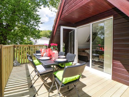 An outdoor wooden deck with a dining table set with plates and cups and six chairs with green cushions at 17 Valley Lodges in Gunnislake
