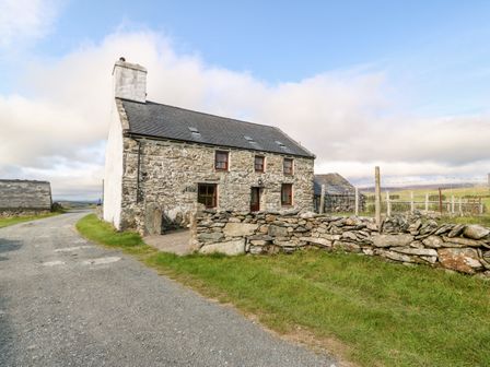A stone house with a slate roof beside a gravel road and a stone wall in a rural area at Ereiniog in Tremadog