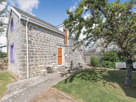 A stone building with wooden doors and purple window frames with outdoor tables and chairs on a path and grass at The Coach House in St Florence
