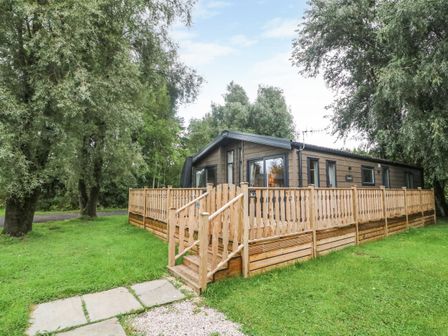 A wooden cabin with a wooden fenced deck surrounded by grass and trees at Coniston View 9 Dock Acres near Tewitfield