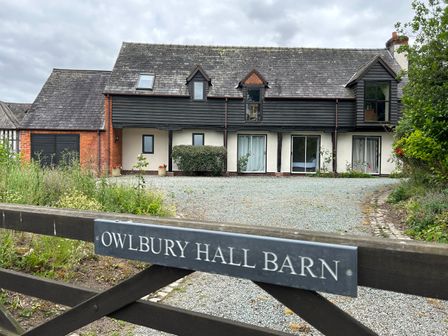 A barn style house with black wooden panels and white walls behind a gate with a sign that says Owlbury Hall Barn in Snead near Bishops Castle Montgomery