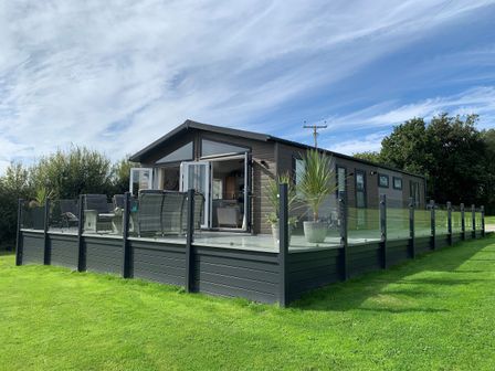 A single-story cabin with a glass and metal railing deck with outdoor furniture and potted plants in a grassy area at Fairview Lodge in Pwllheli