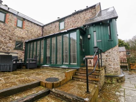 An exterior view of a stone and green wooden building with a patio and outdoor chairs at Skomer Cottage in Sandy Haven near Milford Haven