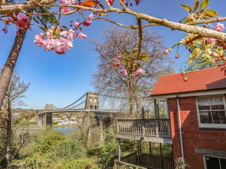 A red brick house with a balcony near a river with a large suspension bridge and trees with pink blossoms at Ceris Lodge Bangor near Menai Bridge