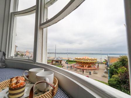 A window seat with two mugs and a muffin on a tray overlooking a carousel and seaside at Shore View in Weymouth