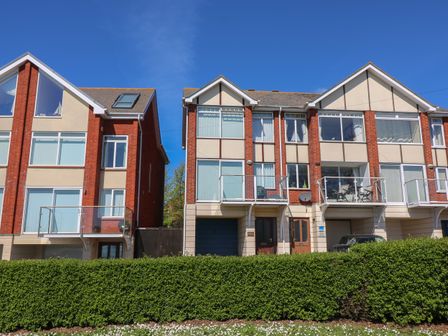 A row of modern brick and beige townhouses with balconies and large windows behind a green hedge at Kiama in Weymouth