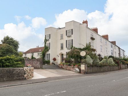 Large white building with multiple windows and chimneys next to a road with stone walls and shrubs at Alcombe House in Minehead