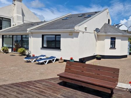 A white house with black window frames two blue sun loungers and a wooden bench on a paved area at High Tide in Duncannon County Wexford