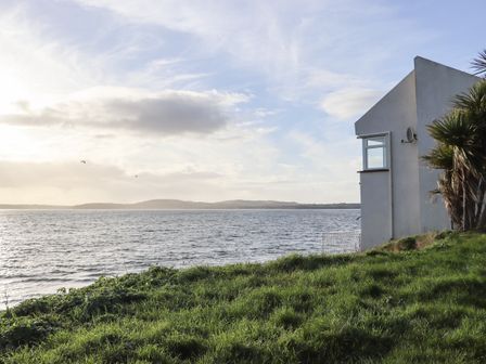 A grassy area next to a body of water with a white building on the right and hills in the distance at Beach House in Duncannon County Wexford