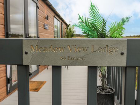 A gate with a Meadow View Lodge sign in front of a wooden lodge with glass doors and a potted plant on the porch at Plot 18 Meadow View in Felton