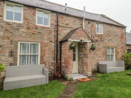 A stone cottage exterior with two gray benches on grassy lawn and a small porch with hanging plants at Donni Hall Cottage in Beadnell