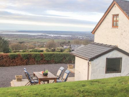 An outdoor patio with a wooden table and chairs next to a small building and overlooking a landscape with fields and water at Beech Tree Cottage in Waunfawr