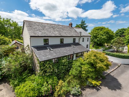 A two story house with a slate roof surrounded by trees and bushes at Waterland Farmhouse in Bradworthy
