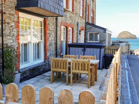 An outdoor patio with a wooden table and chairs next to a stone building by the sea at Salty Sea Dog Trebarwith Strand near Tintagel