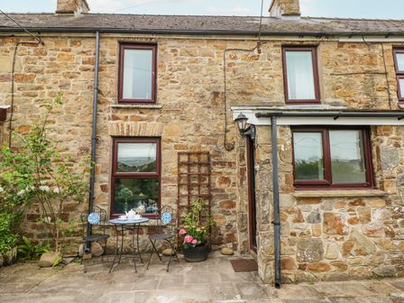 A stone exterior wall of a cottage with three windows a small table with two chairs and potted plants outside Dunnock Cottage Broad Haven