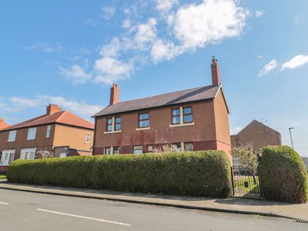 A two-story house with brown walls and two chimneys behind a trimmed hedge and gate on a street at Bisley House in Amble