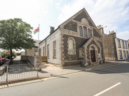 A stone building with arched windows and a flagpole with a flag next to a street at The School House in Holyhead