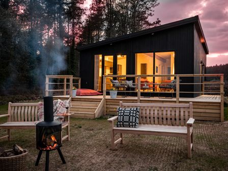A modern cabin with a wooden deck and outdoor seating near a fire pit at Murmur y Coed in Newborough