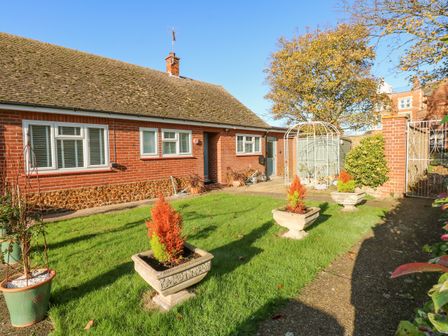 A brick house with white framed windows and a grassy garden with potted plants and a white garden arch at See The Sea in Hunstanton