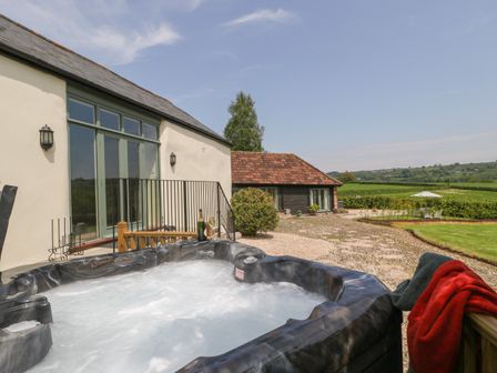 A bubbling outdoor hot tub next to a cream building with large green windows overlooking a gravel yard and countryside at The Hayloft in Stockland Honiton