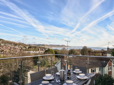 An outdoor balcony with a dining table set with plates bowls glasses and a bottle overlooking houses trees hills and the sea at 5 Victoria House in Lyme Regis