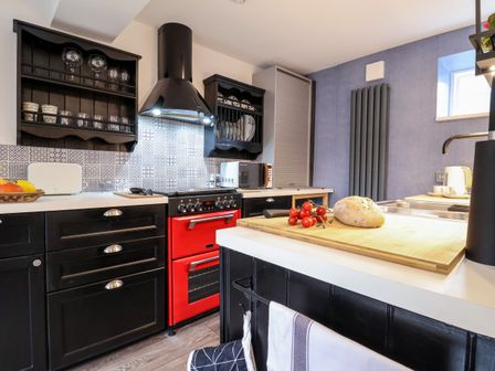 A kitchen with black cabinets a red oven a wooden cutting board with bread and tomatoes and a tiled backsplash at Tower End Cottage in Melton