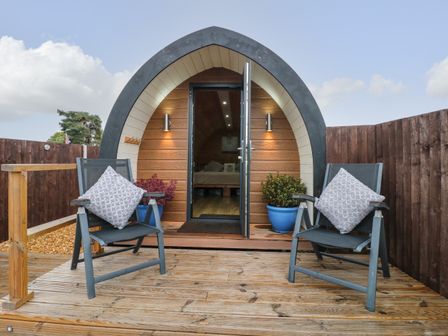 An outdoor wooden deck with two chairs and potted plants in front of an arched tiny house entrance at The Pod at Bank House Farm The Blythe near Hixon