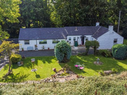 A white long cottage house with a slate roof and garden with lawn chairs and bird feeder at Ty Bach Beaumaris