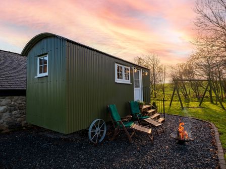 A green corrugated metal hut with a white door and windows with two wooden chairs and a fire pit outside on gravel at The Pleasant Hut at MountPleasant Farm Roosebeck near Ulverston