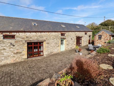 A stone barn with windows and a door next to a patio with outdoor furniture and plants at Great Meadow Barn in Crantock