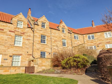 A stone building with multiple windows and a red tiled roof with bushes and a pathway in front at Harebell in Sandsend