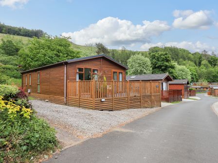 A wooden lodge with a fenced deck next to a paved road with other wooden buildings and trees in the background at Hollytree Lodge Beckside 1 Limefitt Park near Windermere