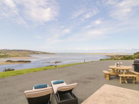 An outdoor patio with two lounge chairs and a wooden picnic table overlooking a river and hills at Upper Killult in Killult near Gortahork County Donegal