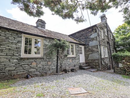 A stone cottage with multiple windows and a white door surrounded by trees at St Giles in Elterwater near Chapel Stile