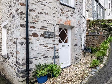 Stone wall building with white door and sign reading Honey Cottage with plants in blue pots and black bench outside at Honey Cottage in Port Isaac
