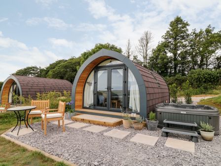 Two arched wooden cabins with glass doors a patio set and a hot tub surrounded by plants at Ash in Saltash