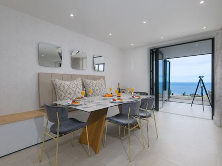 A dining area with a marble table set for breakfast and a view of the sea through open glass doors at Monowai Heights in Carbis Bay