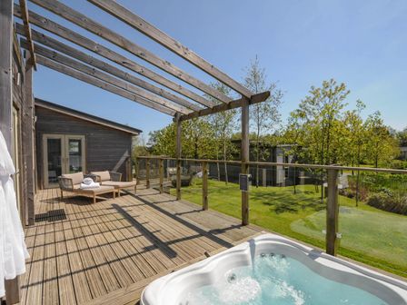 A wooden deck with a hot tub a lounge chair and a wooden pergola overlooking a garden at 11 Meadow Retreat in Dobwalls